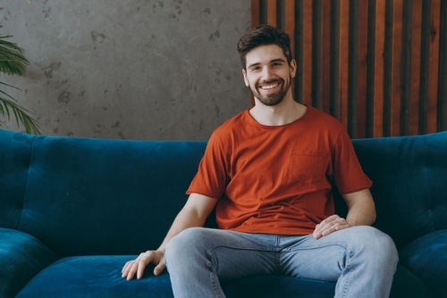 young-smiling-cheerful-man Young adult male smiling while sitting on a dark blue couch inside